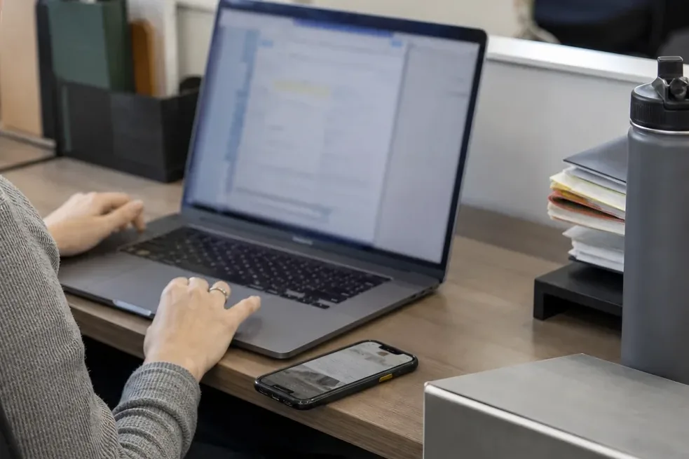 Assistant forwarding receipt emails from a laptop with a phone on the desk beside it